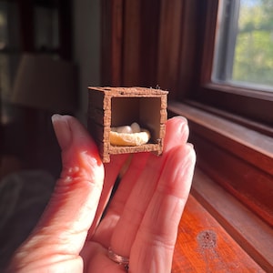 May include: A miniature wooden crate with a nest containing three white eggs. The crate is brown and has a textured appearance, held in a person's hand near a window.
