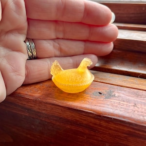 May include: A small, translucent, golden-yellow figurine of a hen sitting on a nest. The object features a textured surface with a detailed comb on the head and a fanned tail. The base is shaped like a round, woven basket with a beaded rim.
