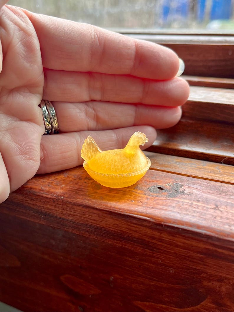 May include: A small, translucent, golden-yellow figurine of a hen sitting on a nest. The object features a textured surface with a detailed comb on the head and a fanned tail. The base is shaped like a round, woven basket with a beaded rim.