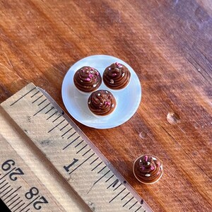May include: Miniature chocolate cupcakes with pink and white sprinkles on a small white plate. A wooden ruler is in the foreground, and a single cupcake is separate from the plate. The cupcakes are on a wooden surface.