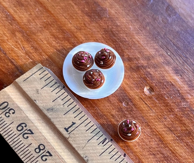 May include: Miniature chocolate cupcakes with pink and white sprinkles on a small white plate. A wooden ruler is in the foreground, and a single cupcake is separate from the plate. The cupcakes are on a wooden surface.
