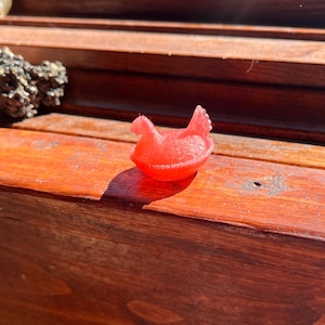 May include: A small, translucent red figurine shaped like a chicken sitting in a nest. The object has a smooth, molded texture and a distinct silhouette of a bird with a raised tail and head, resting within a circular, bowl-like base structure.