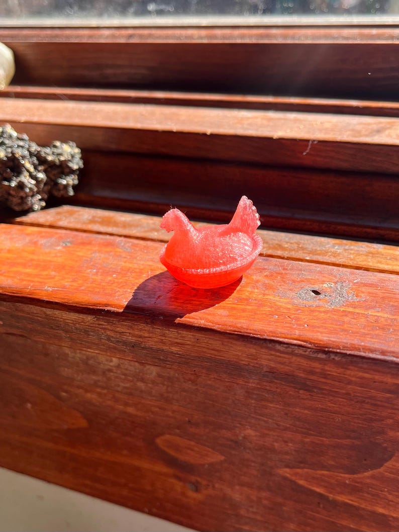 May include: A small, translucent red figurine shaped like a chicken sitting in a nest. The object has a smooth, molded texture and a distinct silhouette of a bird with a raised tail and head, resting within a circular, bowl-like base structure.