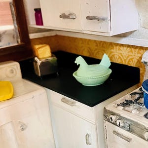 May include: A miniature kitchen scene featuring a white cabinet set with a black countertop. On the counter sits a silver toaster with toast, a pale green ceramic hen-shaped dish, and a blue speckled enamel pot on a white stove.
