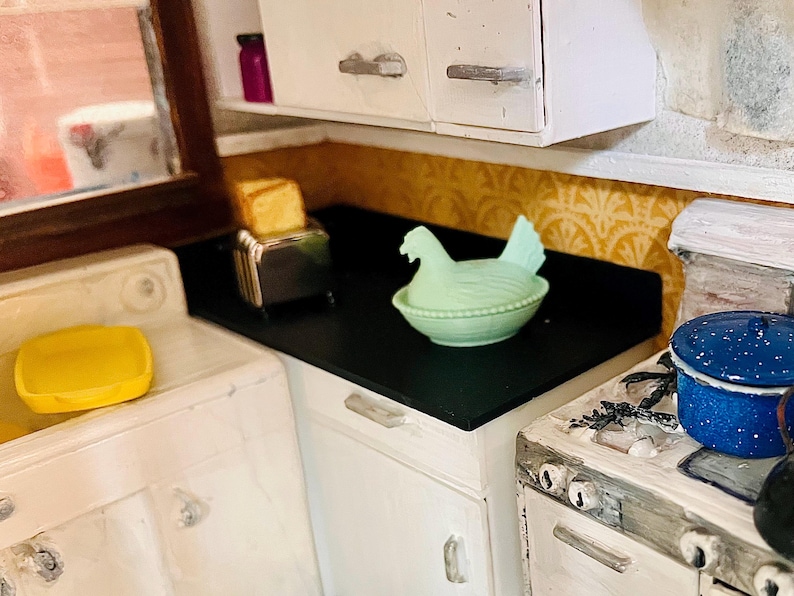 May include: A miniature kitchen scene featuring a white cabinet set with a black countertop. On the counter sits a silver toaster with toast, a pale green ceramic hen-shaped dish, and a blue speckled enamel pot on a white stove.