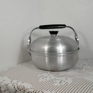 May include: A vintage aluminum cooking pot with a lid and a black handle. The pot has a rounded shape and a small black knob on the lid. It sits on a white lace tablecloth.
