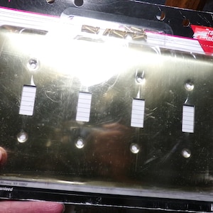 以下が含まれることがあります： 真鍮仕上げの4連電気壁プレートで、ライトスイッチ用の長方形の切り抜きが4つあります。プレートは、透明なプラスチック袋に、白と赤のラベルで「真鍮仕上げ」と表示されています。