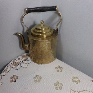 Puede incluir: Una tetera de latón de color dorado con un asa de madera y un diseño floral decorativo. La tetera tiene una tapa con varias capas.