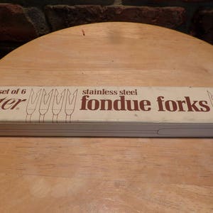 May include: A white box with brown text that reads "set of 6 Oster stainless steel fondue forks". The box shows a drawing of six fondue forks.