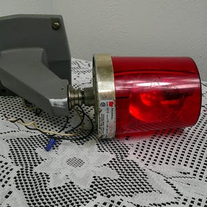 May include: A vintage red emergency light with a grey mounting bracket. The light has a cylindrical red lens and a silver-coloured metal band. Wires and a blue connector are visible. The light is on a white lace tablecloth.