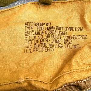 Könnte beinhalten: Ein beigefarbenes Segeltuch-Zubehörset für ein Ein-Mann-Floß. Das Set enthält aufgedruckten Text: "ACCESSORY KIT TYPE I FOR 1 MAN RAFT (TYPE C-2A)", "SPEC. MIL-A-5330A(USAF)", "STOCK NO. AIR FORCE 2010-001200", "DATE OF MFR. JUNE-1953" und "U.S. PROPERTY".