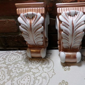 Op de afbeelding: Een paar decoratieve wandbeugels met een witte en gouden afwerking. De beugels hebben een bladontwerp en zijn gemaakt van keramiek.