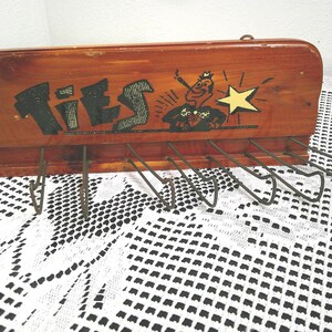 Könnte beinhalten: Ein rechteckiger Krawattenhalter aus Holz mit polierter Oberfläche, der das Wort TIES in schwarzer Schrift und eine grafische Illustration einer Person mit Zigarre neben einem gelben Stern zeigt. Er enthält sechs Metallhaken.