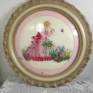 Puede incluir: Una obra de arte en punto de cruz enmarcada que presenta a una mujer con un vestido rosa, sosteniendo una flor y una cesta, con una mariposa. La obra de arte está enmarcada en un marco redondo con un borde dorado ornamentado y un borde interior rosa.