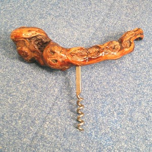 May include: A handcrafted corkscrew with a unique, organic design. The handle is made from a piece of natural wood, showcasing a rich, reddish-brown color and intricate grain patterns. The metal screw and lever are silver-toned, providing a contrast to the wood.