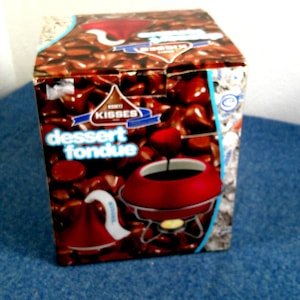 May include: A box of Hershey's Kisses dessert fondue. The box features a design of chocolate kisses and a red fondue pot with the words "dessert fondue" in blue. The box is on a blue surface.