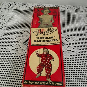 May include: A rectangular red cardboard box for Hazelle's Popular Marionettes. The box features an illustration of a child operating a marionette, a clown marionette in a red and white polka dot suit, and the text "Hazelle's Popular Marionettes".