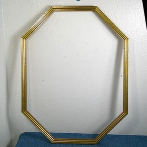 May include: An octagonal gold-colored picture frame. The frame has a ridged design and is empty, ready for a photo or artwork. The frame is set against a white wall and a blue surface.