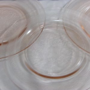 Puede incluir: Tres platos de vidrio rosa con un delicado diseño grabado. Los platos están apilados uno encima del otro, con el plato superior ligeramente desplazado.