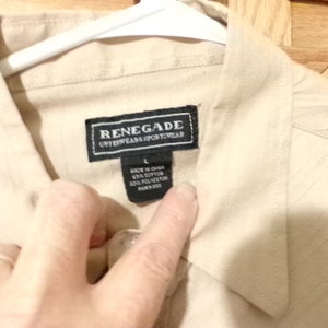 K&ouml;nnte beinhalten: Ein beigefarbenes Hemd mit Kn&ouml;pfen und einem schwarzen Etikett mit der Aufschrift "Renegade Outerwear Sportwear" und der Gr&ouml;&szlig;e "L". Das Etikett tr&auml;gt au&szlig;erdem die Aufschrift "Hergestellt in China 60% Baumwolle 40% Polyester  Importiert."