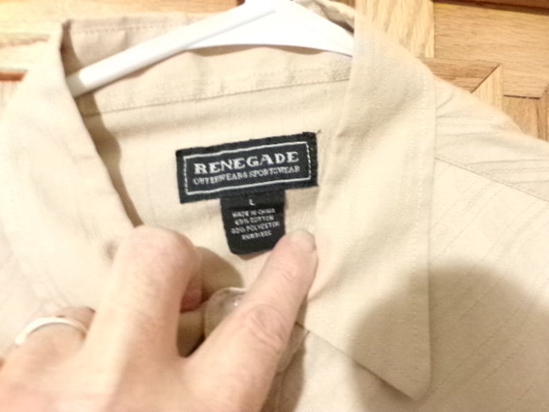 K&ouml;nnte beinhalten: Ein beigefarbenes Hemd mit Kn&ouml;pfen und einem schwarzen Etikett mit der Aufschrift "Renegade Outerwear Sportwear" und der Gr&ouml;&szlig;e "L". Das Etikett tr&auml;gt au&szlig;erdem die Aufschrift "Hergestellt in China 60% Baumwolle 40% Polyester  Importiert."