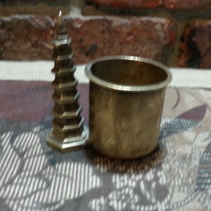 Könnte beinhalten: Eine kleine, messingfarbene Pagoden-Skulptur steht neben einer messingfarbenen zylindrischen Tasse. Die Pagode hat mehrere Ebenen und eine spitze Spitze. Die Tasse hat ein einfaches, eingraviertes Design.