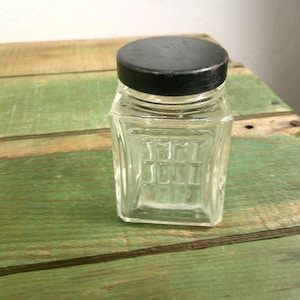 Peut inclure: Un petit bocal en verre transparent de forme carrée présentant un motif en grille texturé sur ses côtés. Le récipient est surmonté d'un couvercle métallique noir, plat et circulaire, montrant des signes d'usure sur les bords.
