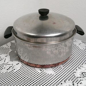 以下が含まれることがあります： 蓋と黒い取っ手が付いたステンレス製の鍋。鍋底は銅色で、蓋には黒いノブが付いています。白いレースのテーブルクロスの上に置かれています。