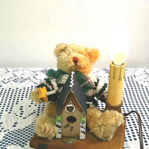May include: A teddy bear lamp with a birdhouse and candle design. The bear is wearing a green and white scarf and holding a yellow star. The lamp has a wooden base and a warm, glowing light.
