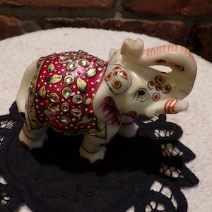May include: White marble elephant figurine with red, gold, and green painted details and rhinestones. The elephant has a raised trunk and is decorated with a floral pattern.