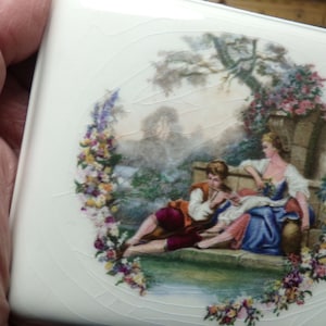 以下が含まれることがあります： 花柄のデザインと、庭のベンチに座っている2人の人物を描いた絵が施された白い陶器製の小物入れです。