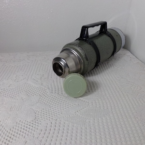 May include: A vintage green metal thermos with a black handle and a green plastic lid. The thermos is lying on a white crocheted surface.