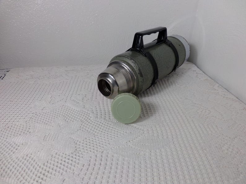 May include: A vintage green metal thermos with a black handle and a green plastic lid. The thermos is lying on a white crocheted surface.