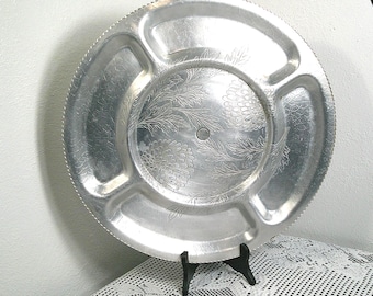 May include: A round, silver-colored serving tray with four compartments. The tray has a decorative floral pattern in the center and along the edges. The rim has a textured, scalloped design. The tray is made of metal.