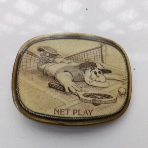 Puede incluir: Una hebilla de cinturón de latón con una ilustración de dibujos animados de una persona jugando al tenis. La hebilla tiene las palabras "NET PLAY" inscritas en ella.