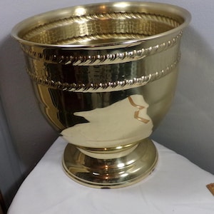 Puede incluir: Un macetero grande de metal dorado con un acabado martillado y un diseño de cuerda alrededor del borde. El macetero tiene una base ancha y un cuerpo redondeado.