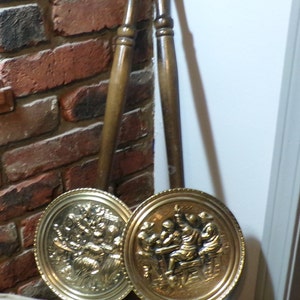 May include: Two antique brass wall plaques with raised relief designs of people in a tavern setting. The plaques are attached to wooden handles.