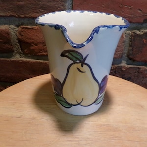 Puede incluir: Una jarra de cerámica blanca con un borde azul y un diseño de pera. La pera es amarilla con hojas verdes y un tallo marrón. La jarra tiene un diseño floral azul y morado alrededor de la base.