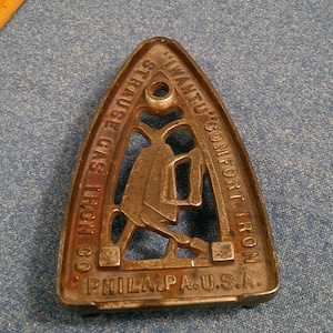 Puede incluir: Antigua plancha de ropa de hierro fundido con el texto "STRAUSE GAS IRON CO." y "WANTU'S COMFORT IRON" y "PHILA. PA. U.S.A." La plancha es triangular con un recorte en forma de asa y una figura de una persona planchando.