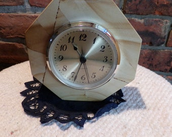 Könnte beinhalten: Eine kleine, dekorative Uhr mit silbernem Zifferblatt und einem hellen, geometrischen Holzrahmen. Die Uhr steht auf einer schwarzen Spitzendeckchen. Der Hintergrund zeigt eine rote Backsteinmauer.