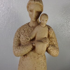 May include: A beige ceramic sculpture of a mother holding a child. The sculpture is detailed with a textured surface.