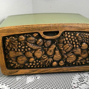 Può includere: Un portapane vintage con coperchio verde chiaro e cornice in legno. Il fronte è intagliato in modo intricato con un rilievo di vari frutti e verdure. Il portapane è su una tovaglia di pizzo bianco. È visibile una maniglia.
