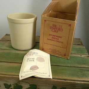 Puede incluir: Un recipiente de gres beige para hornear pan de The Pampered Chef, acompañado de su caja de cartón original y un folleto de recetas e instrucciones. La caja presenta texto en rojo que identifica el producto y la empresa.