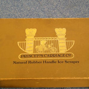 May include: A rectangular, gold-colored box with the text "PRINCETON CARRIAGE CO" and "Natural Rubber Handle Ice Scraper" in black. The box features a gold illustration of a carriage and gate.