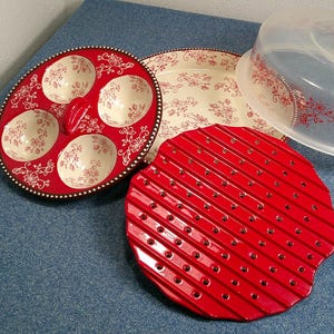 May include: A collection of serving dishes and a clear plastic lid. The red egg serving dish has six egg-shaped indentations with floral designs. A red, ridged serving dish and a floral patterned plate are also present.