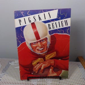May include: Vintage "Pigskin Review" magazine cover featuring a football player in a red uniform and white helmet with a red stripe. The background has blue and white accents. The magazine is displayed on a black stand.