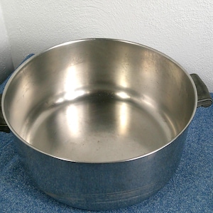 May include: A large, stainless steel cooking pot with two black handles. The pot has a wide, open design and a shiny interior. It sits on a blue surface, with a white wall in the background. The pot is designed for cooking.