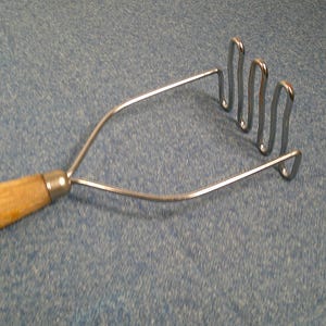 May include: A potato masher with a wooden handle and a stainless steel mashing head. The handle is light brown, and the mashing head is made of looped wires. The masher is designed for kitchen use.