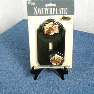 Pode incluir: Uma placa de interruptor de madeira preta com detalhes decorativos de instrumentos musicais. A placa do interruptor é embalada em um cartão branco com a palavra "SWITCHPLATE" no topo. Um pequeno parafuso está incluído. A placa do interruptor é exibida em um suporte preto.