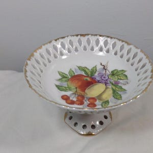 Puede incluir: Cuenco de fruta de porcelana blanca con un borde dorado y un delicado diseño calado. El cuenco presenta un diseño pintado a mano de fruta, incluyendo manzanas, uvas y cerezas.